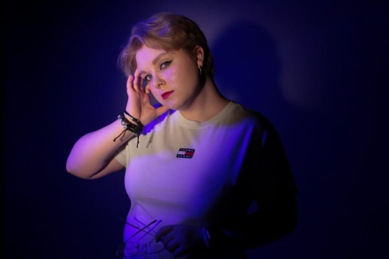 Woman on white shirt looks at the camera, while purple and pink lights reflect on her back.