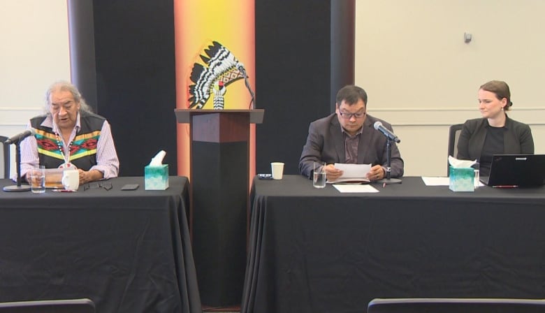 Three people sit at a long table in a conference room.