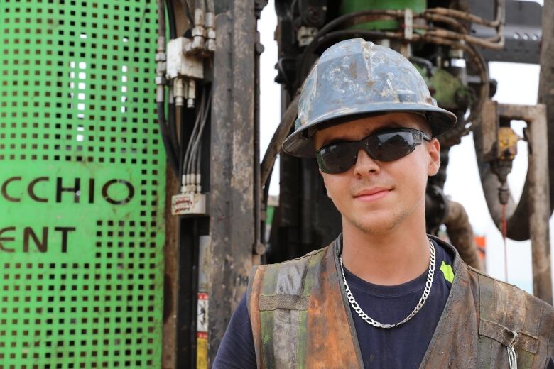 Young man in hard hat and sunglasses in front of some muddy heavy equipment with pipes and wires.