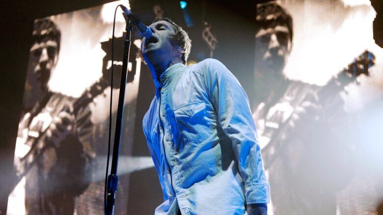 A man sings into a microphone in front of tall screens showing another man playing guitar. 