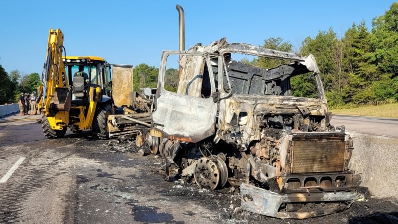 A transport truck cab heavily damaged by fire on the side of a highway on a summer day. There's heavy construction equipment behind it.