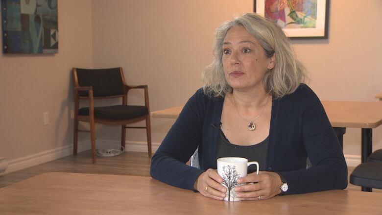 Woman with shoulder length hair in dark sweatshirt at a wooden table, holding a mug