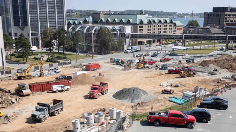 Piles of soil are dotted around the construction site with various pieces of machinery and trucks. A hotel and view of the harbour are in the background