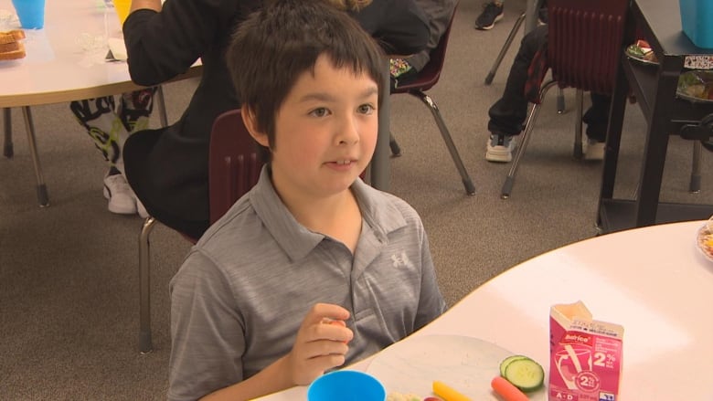 A boy in a grey polo shirt sits in his chair and munches on his vegetables.