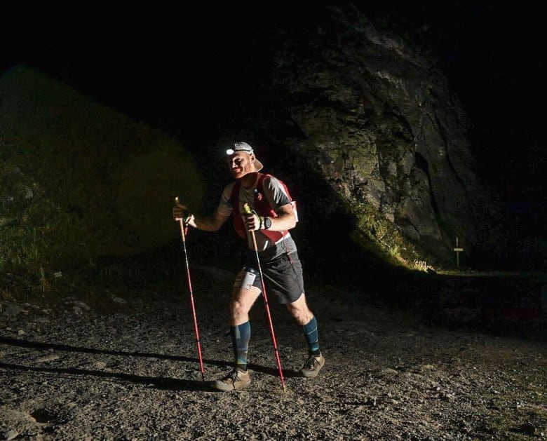 A man is wearing hiking gear and a headlamp on a mountain trail at night.