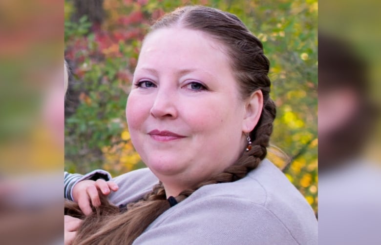 A woman with long hair in braids and wearing a lavender coloured sweater sits outside with trees in fall colours.