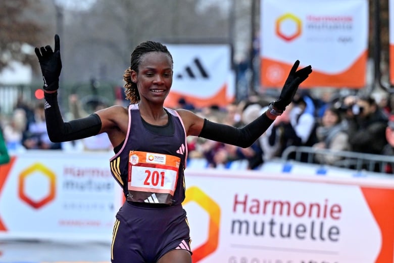 A woman in a purple running suit and black gloves raises her hands in the air and smiles as she crosses the finish line