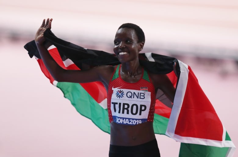 A woman with short hair smiles brightly and waves with a red, green and white flag draped around her shoulers.