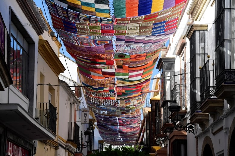 Colourful crocheted blankets drape over a Spanish street, providing shade below. 