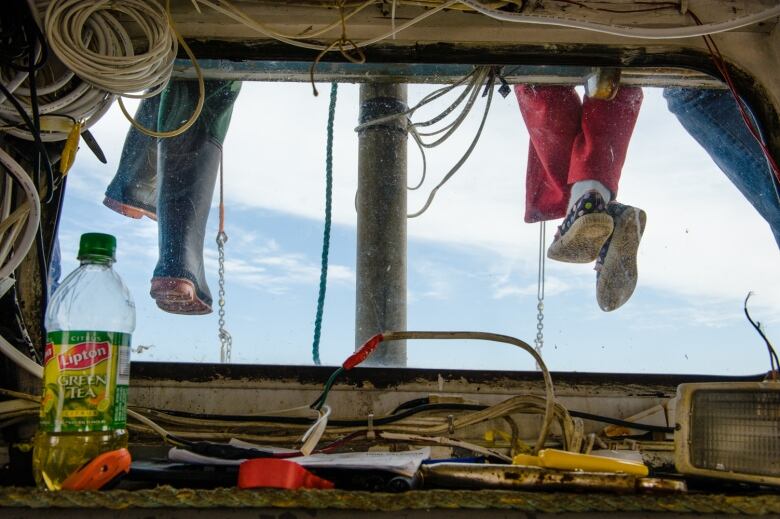 A view from the captain's point of view in a fishing boat, with two sets of legs dangling down in front of the windscreen - one in rubber boots, one in sneakers.