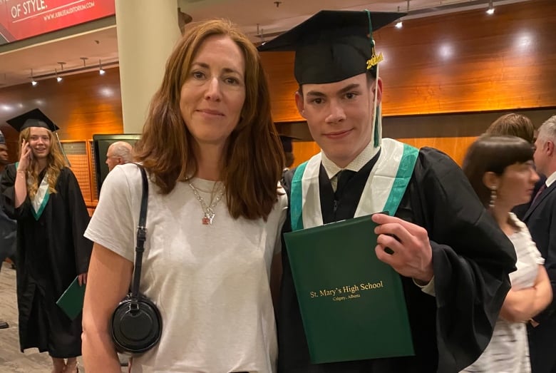 A young man wearing a graduation gown holds a certificate and stands beside a woman. 