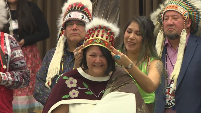 A woman smiles as another woman places a feathered headdress on her head.