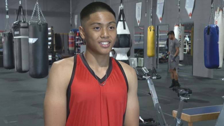 A boy wearing a red sleeveless shirt stands at a boxing gym.