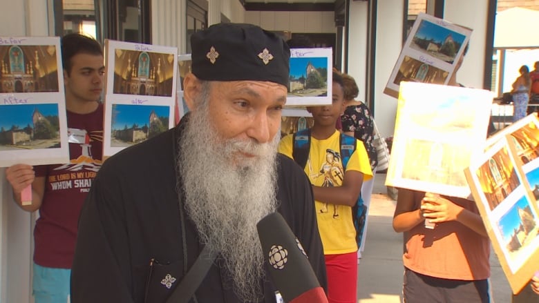 A church priest in a black gown and a cap with a long white beard standing in front of a bunch of children holding signs.