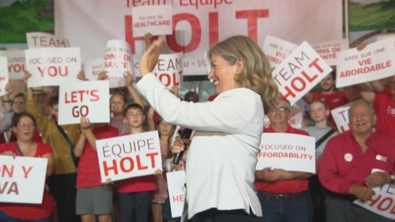 A woman waves to the side of the room, with rows of adults and children wearing red in the background. 