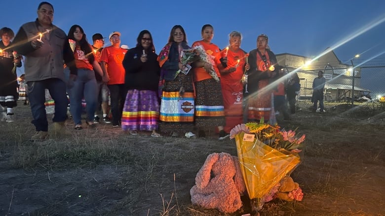 A group of people holding lit candles stand outside in front of a memorial.