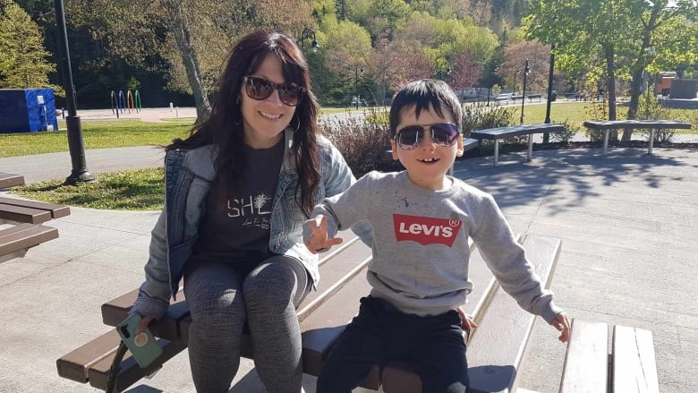 A woman in sunglasses and fark shirt and jacket sitting at a picnic table next to a young boy also wearing sunglasses and grey shirt, smiling. 