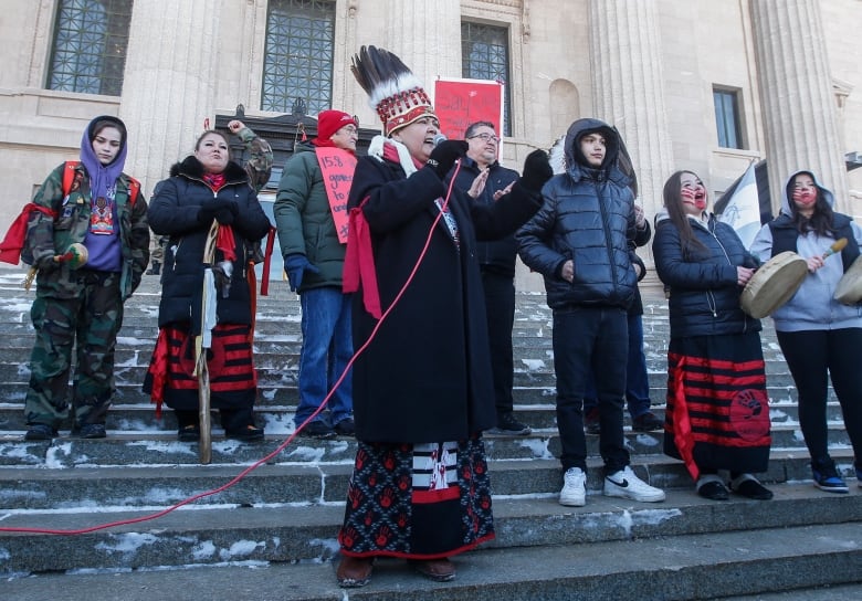 A woman in a traditional First Nations headdress stands on steps and speaks into a microphone.