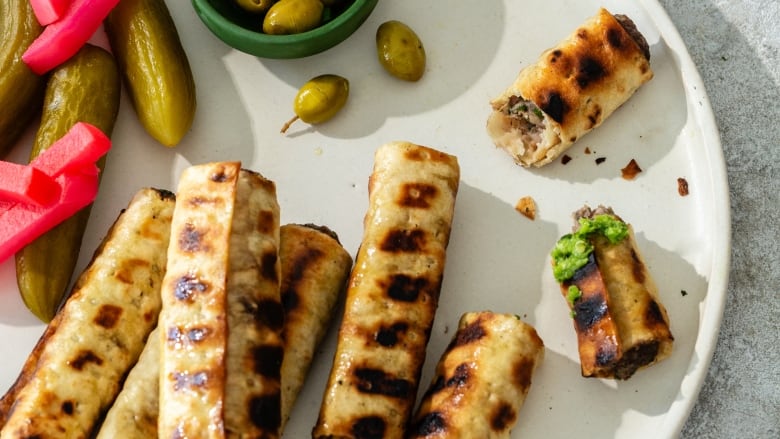 Closeup on a platter of grilled pitas stuffed with kebabs. Green olives and assorted pickles sit next to them. 