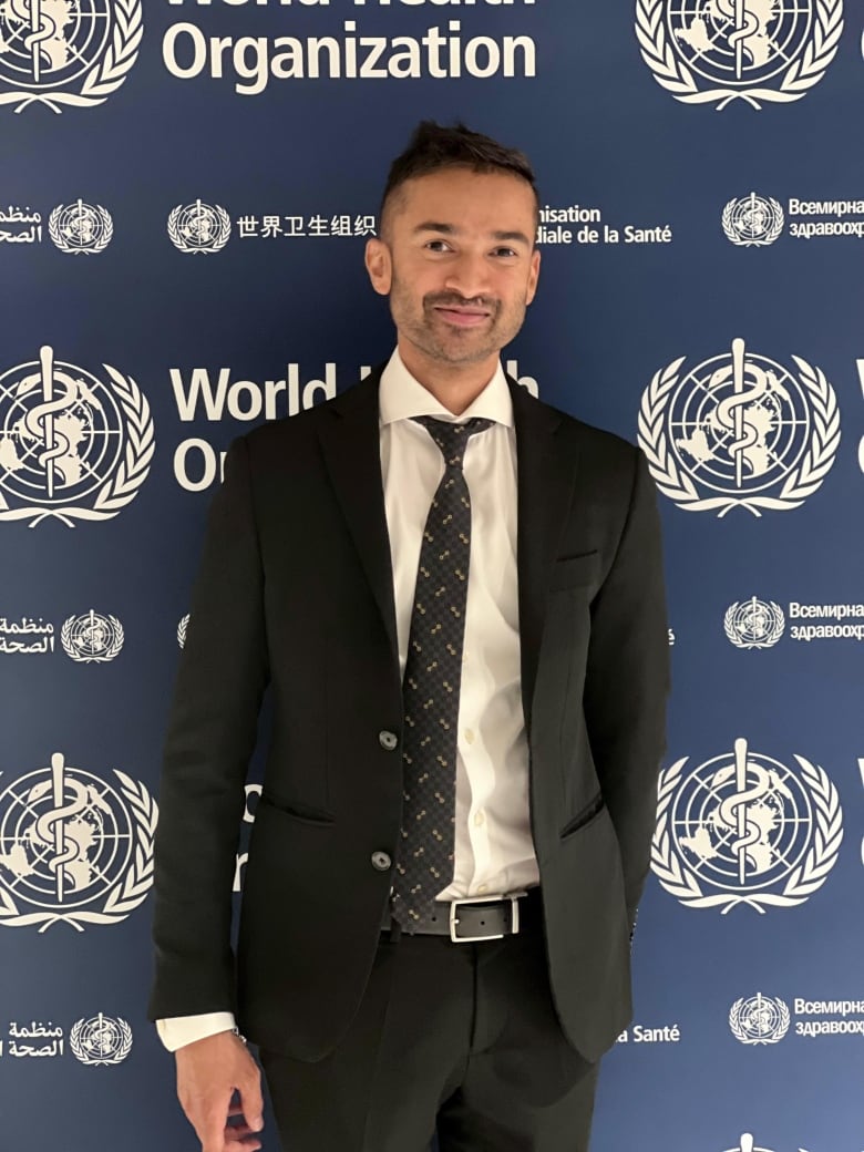 Man in a dark suit and tie standing in front of World Health Organization backdrop. 