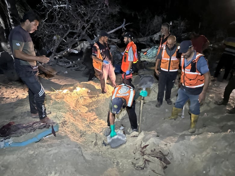 Men in orange vests dig in the sand with shovels at night.