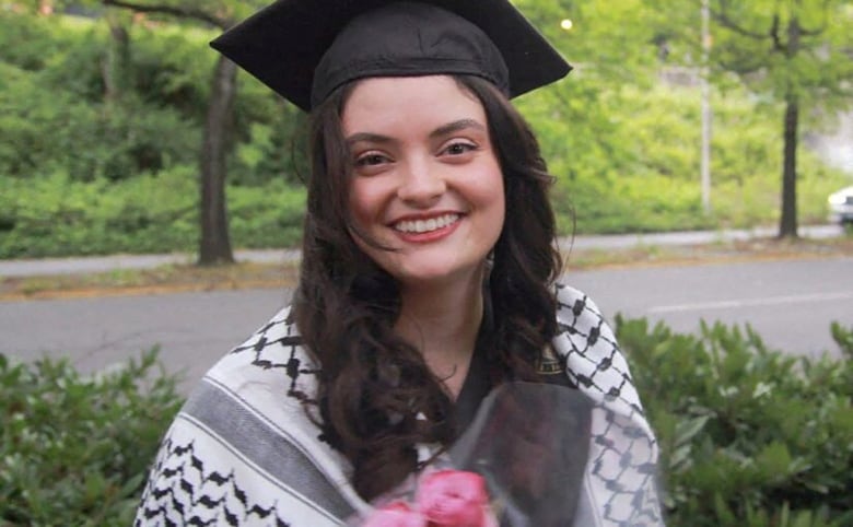 A woman with dark hair wearing a graduation gown and cap while holding a bouquet of pink flowers is shown.