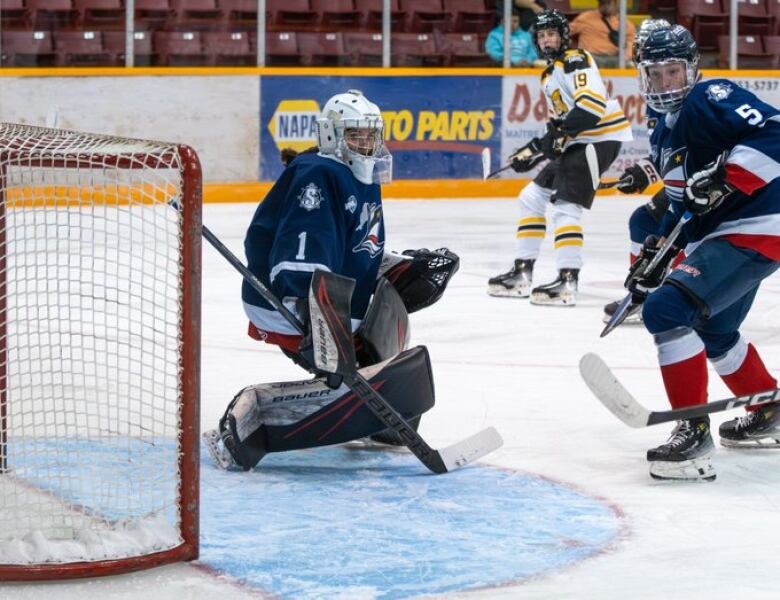 A goalie wearing a navy jersey crouches down in front of a hockey net. Two other hockey players on opposing teams skate nearby.