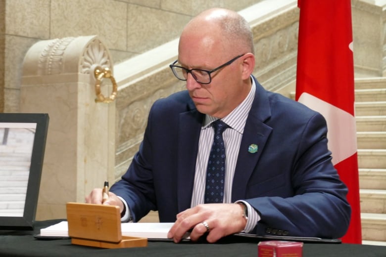 A bald man in a suit sits at a table and signs the page of a book