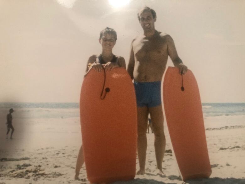 A young girl and her father in swimsuits stand on the beach holding boogie boards.