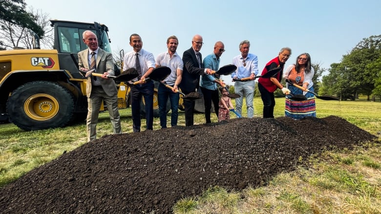 A group of people stand over a mound of dirt with shovels during a community announcement about a recovery centre that will be built soon. 