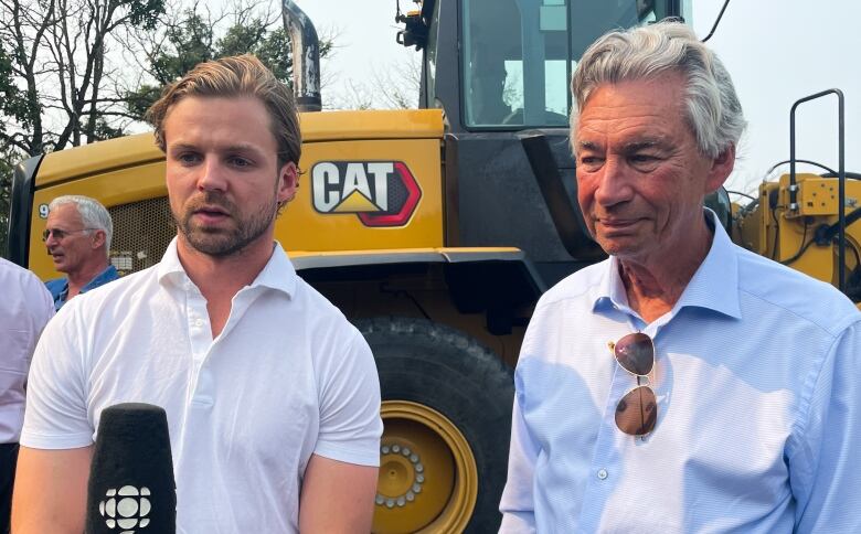 Two men stand before a microphone in front of an excavator in a field. 