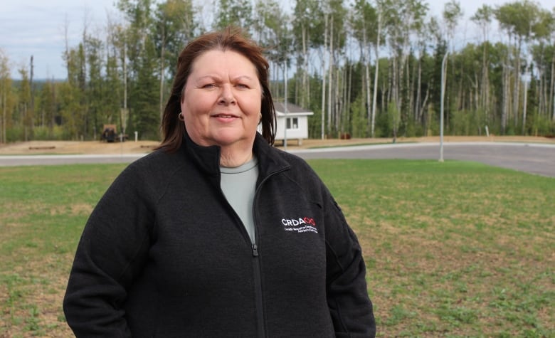 A woman with shoulder-length brown hair and a black zip-up sweater stands in front of a pre-built home in a new subdivision in Conklin, Alta.