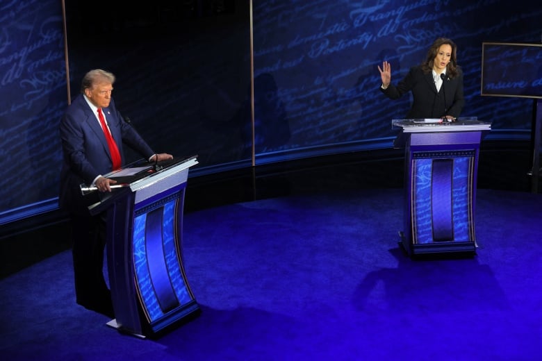 A man and a woman in suits stand behind podiums on a stage lit up in blue.