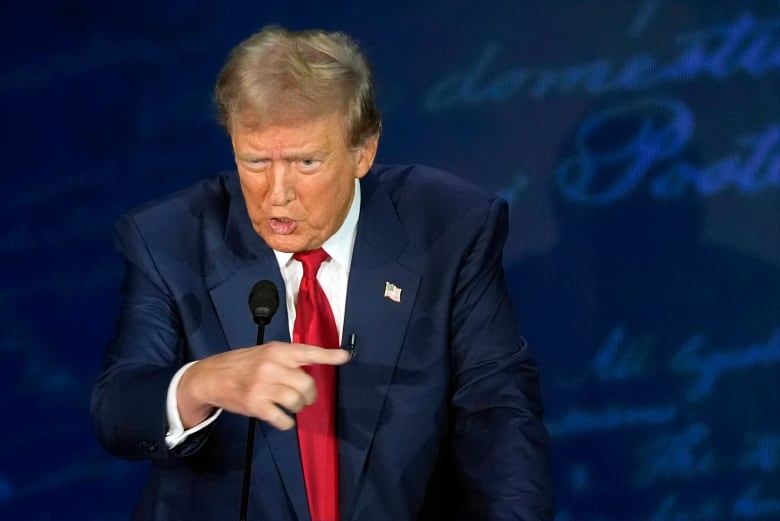 Donald Trump, in a navy blue suit, white shirt and red tie, speaks into a microphone while point to the right. 