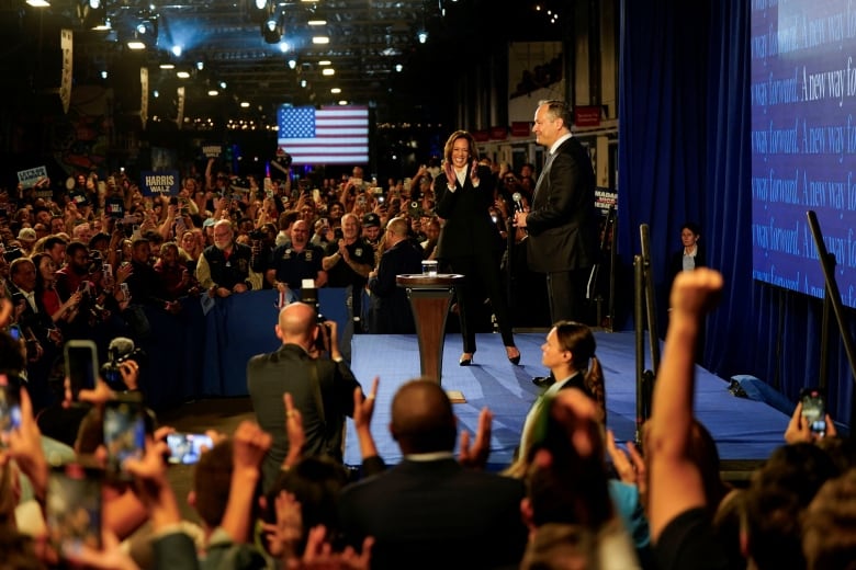 A woman in a suit smiles and claps on stage as a crowd cheers and waves American flags.