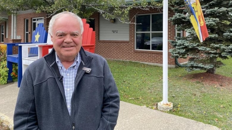 A smiling man with grey hair standing in front of three Adirondack chairs painted like the Acadian flag.