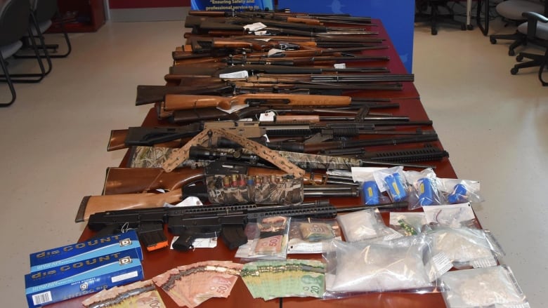 A table with numerous long guns, money and plastic bags full of a white substance is shown.