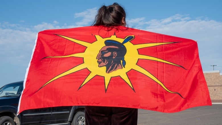 A person is seen from behind standing outside. They are holding a large red flag featuring a person's face against a yellow sun.