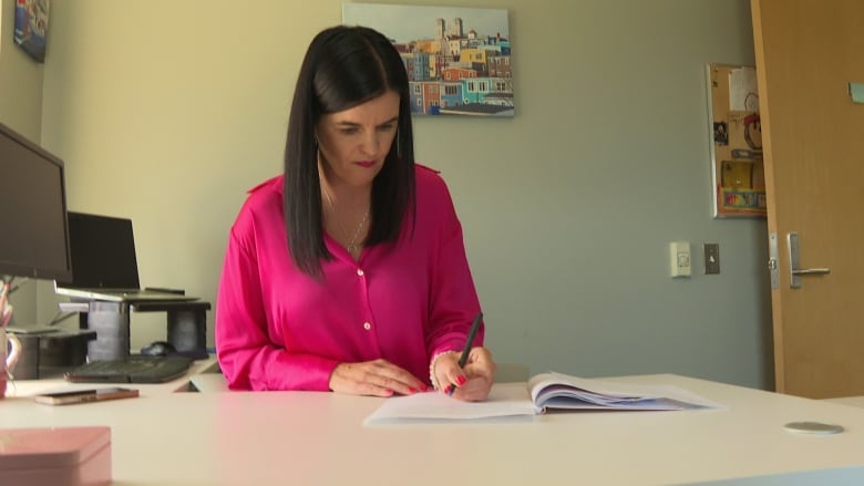 A woman wearing a pink blouse writes at a desk.