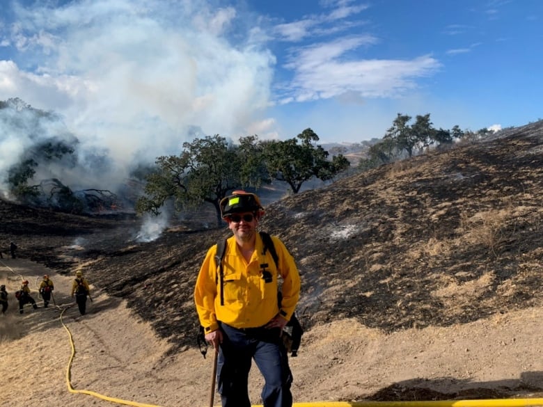 Man in yellow jacket and red helmet stands with smoky landscape in the background