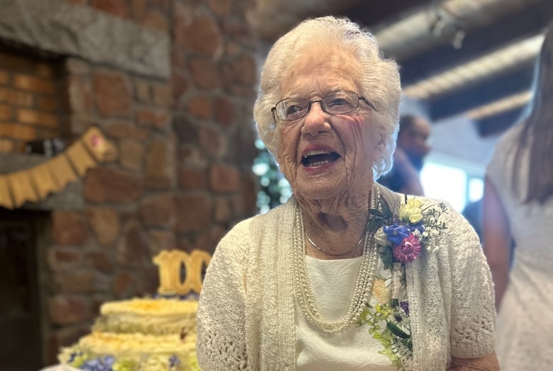 A woman with white hair wearing a dress with a corsage of flowers pinned to it clasps her hands and smiles. Behind her there is a cake with 100 on top of it.