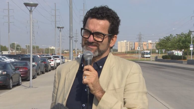 A man in dark hair and glasses holds a microphone and speaks outside as transit buses pass by in the background.