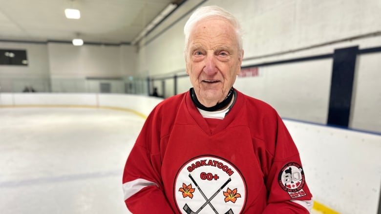 A smiling, elderly man with white hair wearing a red hockey jersey standing on the ice surface in a hockey arena.