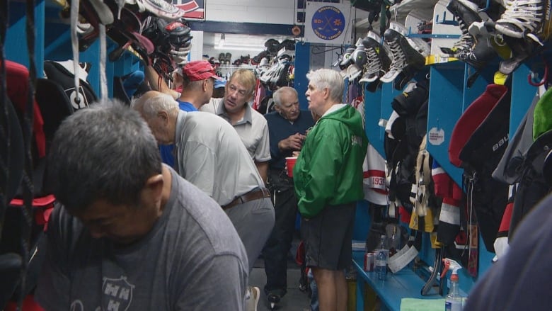 After a game, older hockey players get dressed and have conversations in a dressing room with blue stalls and hanging hockey equipment.