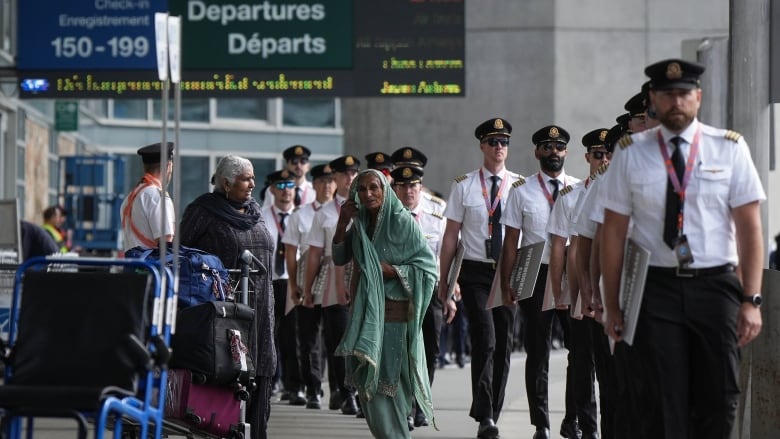 More than a dozen pilots in uniform walk past two passengers pushing a baggage cart as they arrive for an informational picket at an airport.