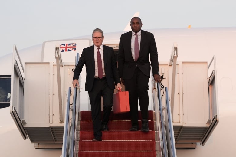 Two clean shaven men in suits, one Caucasian and one dark-complected, are shown at the top of the stairway extending from an aircraft on a tarmac.