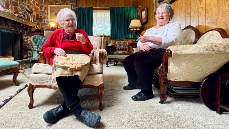 Two women are shown sitting in a living room, one on a chair, the other on a couch.