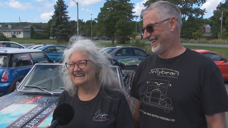 Woman with long grey hair and man with sunglasses and grey hair in matching tee shirts stand next to convertible.