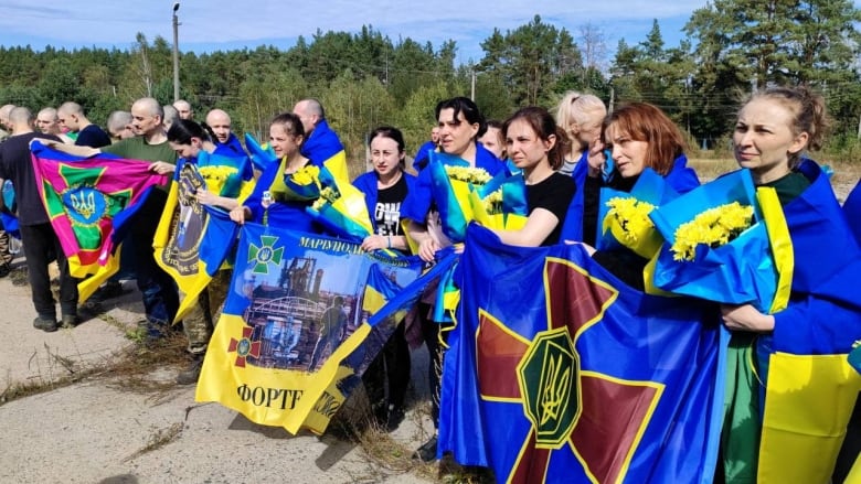 A group of women, holding bouquets of yellow flowers, are wrapped in yellow and blue banners as they stand outside.