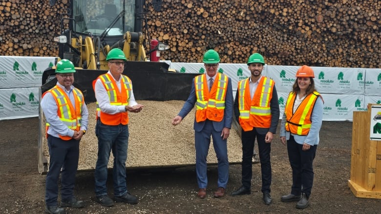 A group of people wearing green hard hats and orange safety vests pose for a photo in front of a loader with a bucket full of wood pellets.